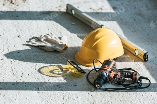 Preview: close up view of hardhat, spirit level, drill and protective gloves with googles at construction