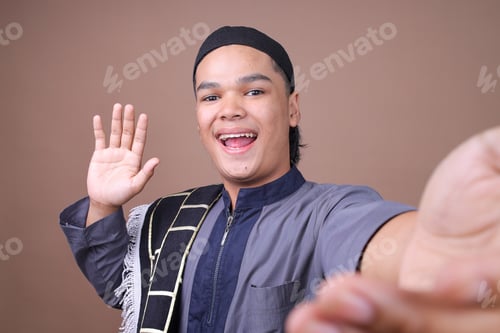 Preview: POV Selfie Of Attractive Young Muslim Man Waving And And Smiling At Camera On Beige Studio Wall