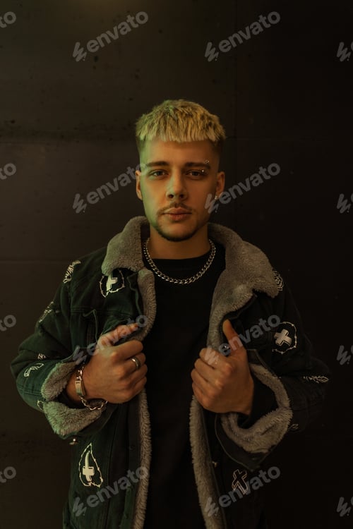 Preview: Fashionable hipster man with hairstyle in stylish black jacket stands near a black wall indoors