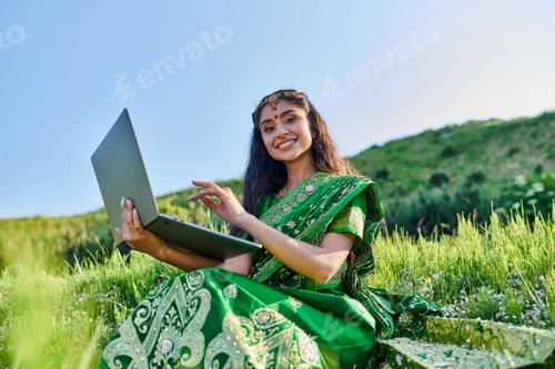 Preview: positive young indian woman in sari looking at camera and holding laptop on grassy field