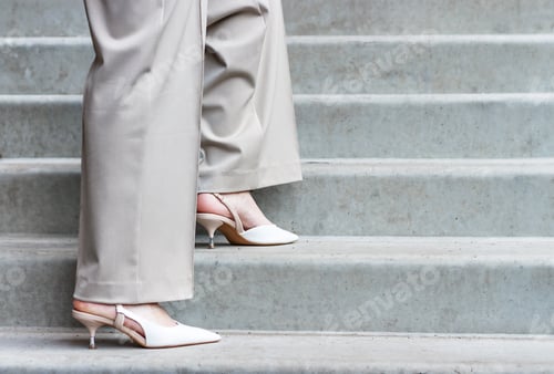 Preview: Side view of female legs in gray trousers and pointed sandals against background of stairs.