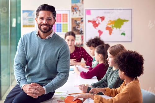 Preview: Portrait Of Smiling Male Elementary School Teacher Working At Desk In Classroom With Students