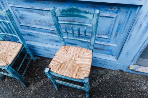 Preview: Two empty rustic blue chairs with straw seats leaning against a blue wall