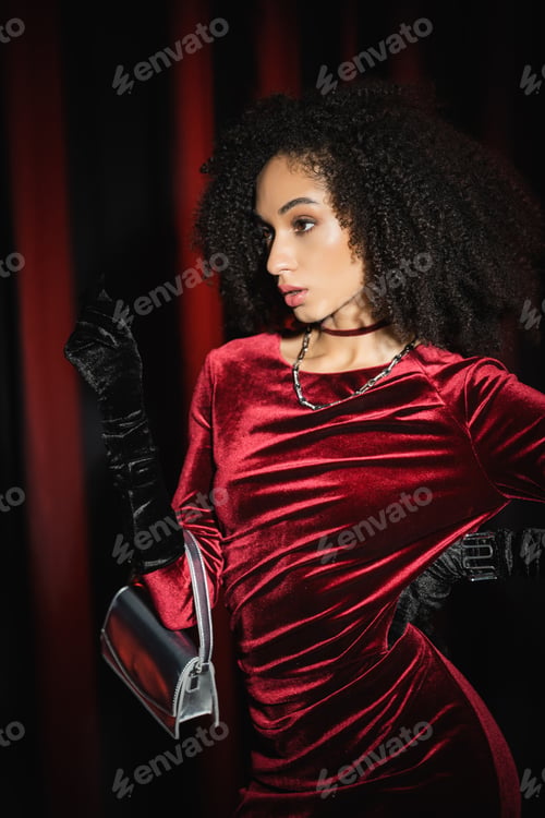 Preview: Portrait of african american woman in elegant dress posing with handbag on burgundy drapery