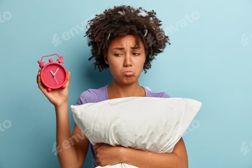 Preview: Photo of sad Afro American young woman purses lower lip, carries pillow, holds alarm clock, unhappy