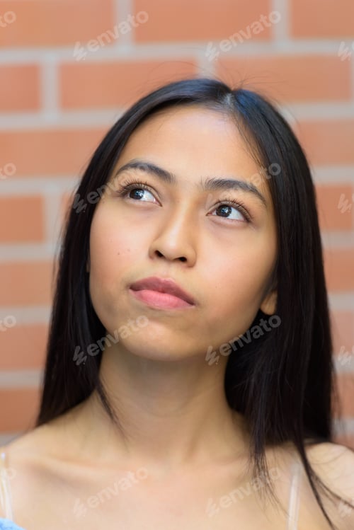 Preview: Face of young beautiful Asian teenage girl thinking while looking up against brick wall