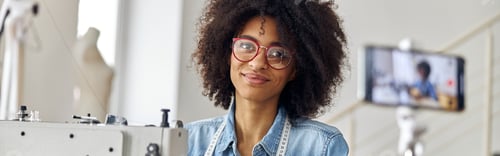 Preview: Pretty African-American woman blogger with scissors and glasses at workplace in sewing workshop