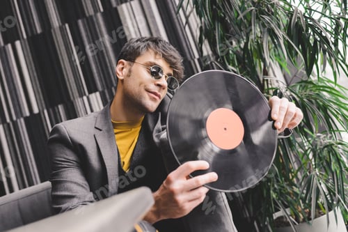 Preview: stylish young man in sunglasses looking at vinyl record