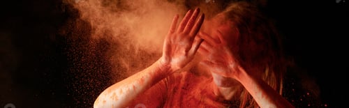 Preview: woman covering face from orange colorful holi paint cloud on black background, panoramic shot