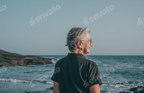 Preview: Rear view of mature smiling senior woman standing at the beach at sunset looking at the horizon