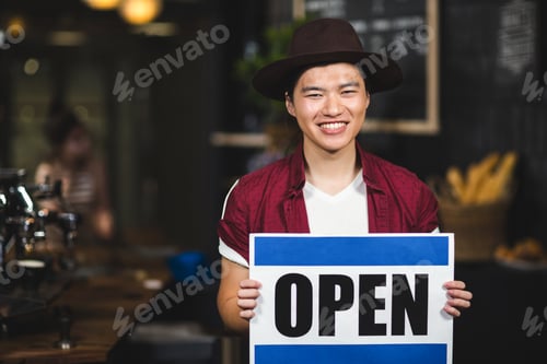 Preview: Smiling Asian man holding Open sign in cozy coffee shop