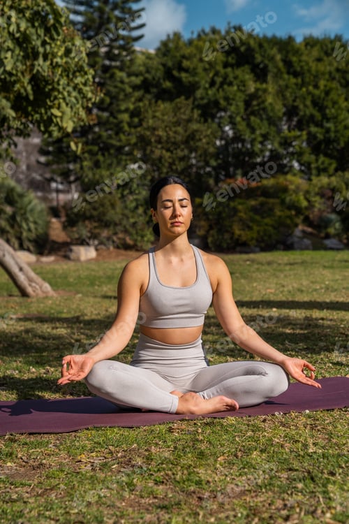 Preview: Vertical shot of a brunette Caucasian woman sitting on a yoga mat in the park doing a meditation