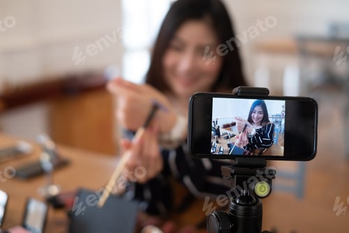 Preview: Young asian woman blogger with makeup cosmetics recording video clip online by smartphone at home.