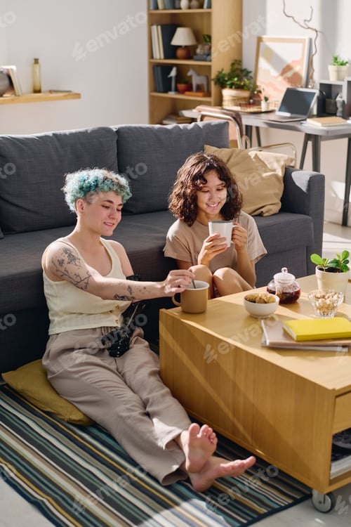 Preview: Two happy young women sitting on the floor by couch and having tea