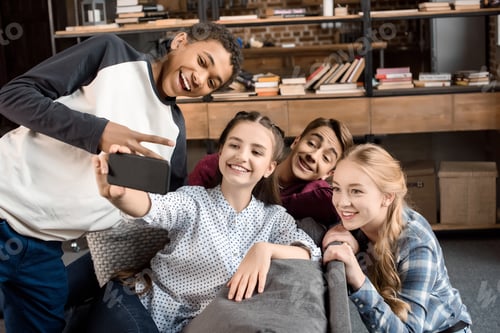 Preview: happy multicultural teenagers group taking selfie on smartphone and sitting on sofa at home,