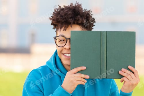 Preview: Playful student hiding behind book outdoors