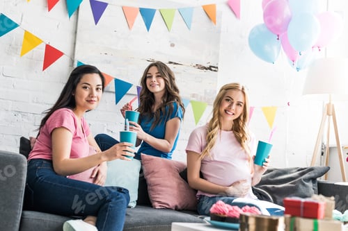 Preview: multicultural friends and pregnant woman holding drinks and looking at camera at baby-party