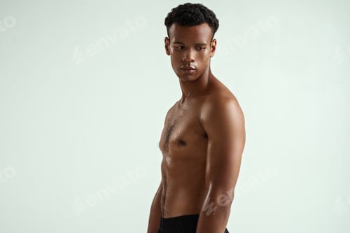 Preview: Side view of young shirtless muscular african man looking away while standing against grey