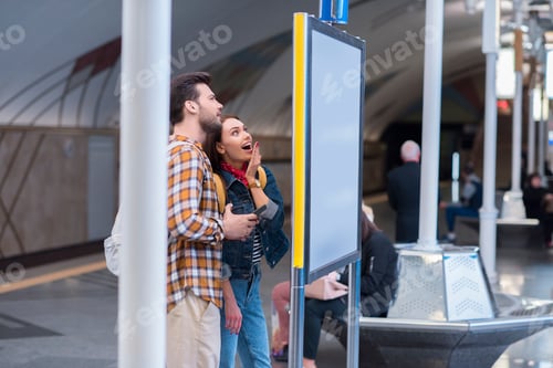 Preview: side view of surprised female tourist with boyfriend looking at information board at subway station