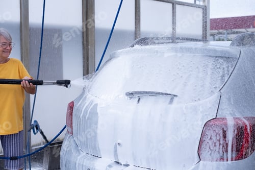 Preview: Smiling senior woman washing her car in a self-service car wash station using high pressure water