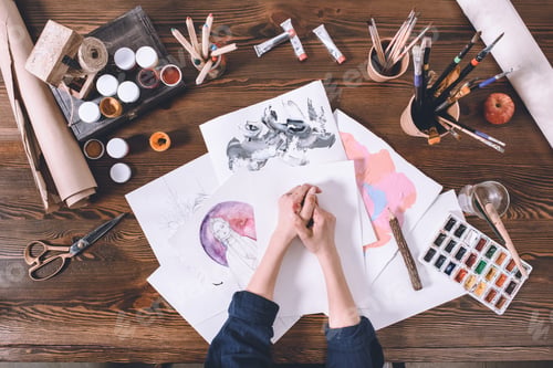 Preview: partial top view of female hands and sketches with paints at artist workplace