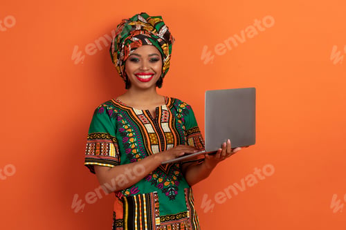 Preview: Happy young african lady using laptop on orange background