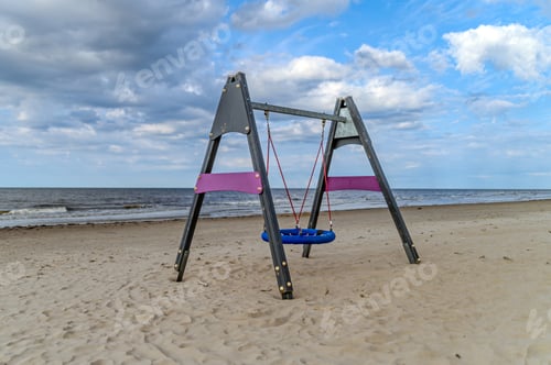 Preview: Colorful swing set on a sandy beach near calm waves under a partly cloudy sky during sunset