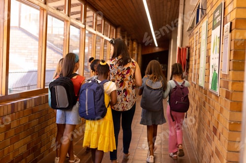 Preview: Diverse female teacher and schoolgirls with bags walking in elementary school corridor