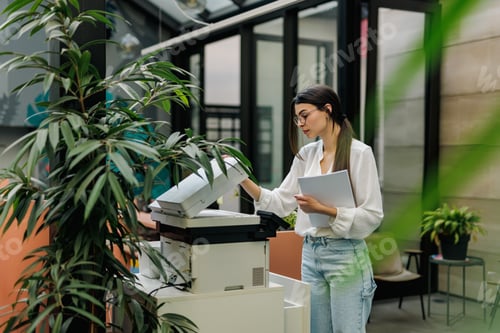Preview: An adult girl working at the office, standing next to the scanner, holding some documents.