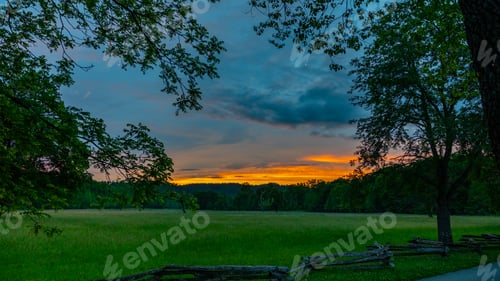 Preview: Sunset over a grassy field with trees and a fence beside the street