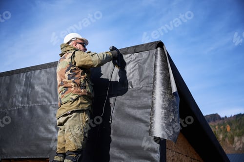 Preview: Male builder installing waterproof membrane on wall of future wooden frame house.