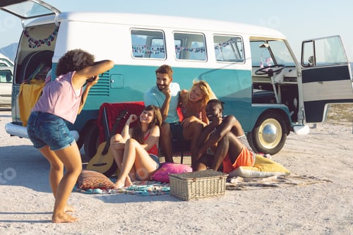 Preview: Young Caucasian woman taking pictures of her friends with digital camera near camper van at beach