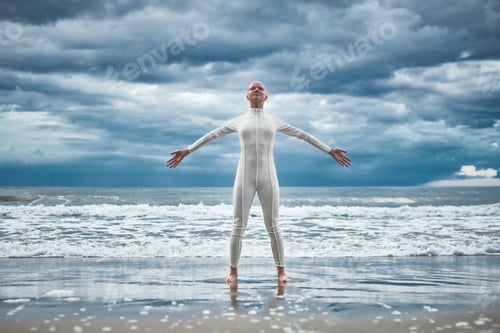 Preview: Happy hairless girl with alopecia in white futuristic suit stands on beach bathed by ocean waves