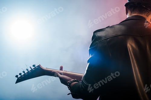 Preview: rear view of male rock star in leather jacket performing on electric guitar on stage with smoke and