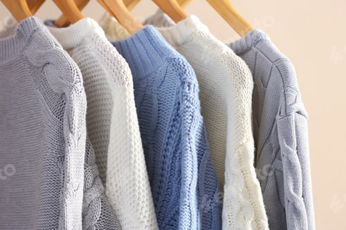 Preview: Collection of warm sweaters hanging on rack near light wall, closeup
