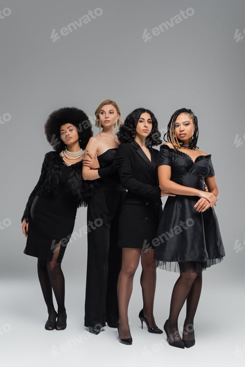 Preview: full length of graceful multicultural girlfriends in black elegant clothing standing on grey