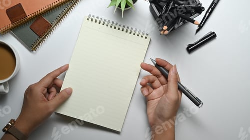 Preview: From above view of female hand holding pen writing in spiral notepad at white working desk.