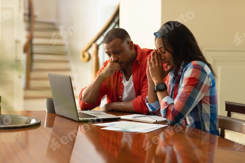 Preview: African american couple looking at laptop while sitting with financial bills at home