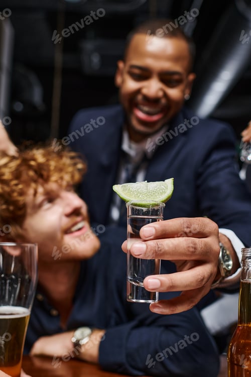 Preview: focus on tequila shot with sliced lime on top, african american man holding drink near friend in bar