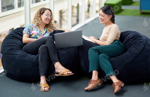Preview: Shot of two businesswomen using a laptop together while sitting on beanbags in an office