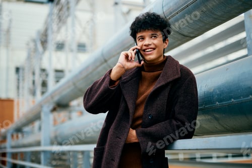 Preview: Young man in brown coat talking on cell phone while standing on metal railing in front of industrial