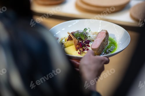 Preview: Back view closeup of a person holding a plate of roasted lamb rump served with sauce and greens