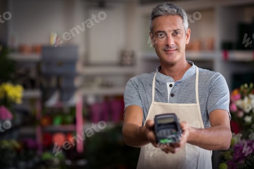 Preview: Middle-aged man wearing apron at counter presenting card terminal with floral displays, copy space