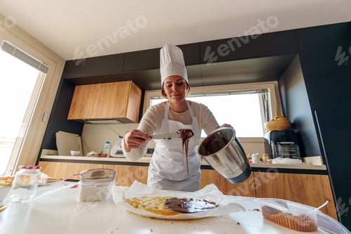 Preview: Woman pastry chef pouring melted chocolate cream on cake base using a blender, in a modern kitchen