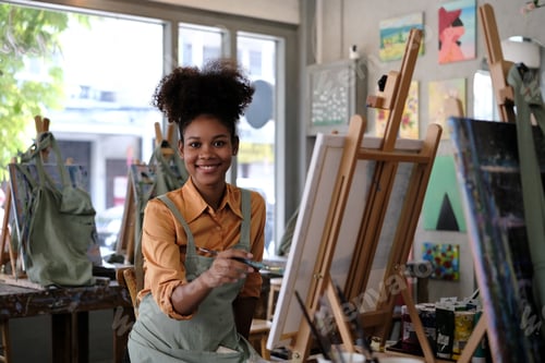 Preview: Portrait of smiling young woman with Afro haired painting picture with brush on easel in art studio.