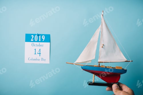 Preview: cropped view of man holding crafted ship near paper calendar with Columbus Day inscription isolated