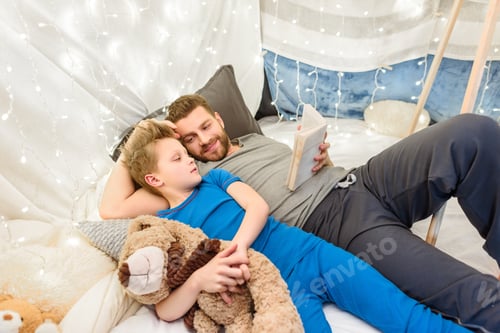 Preview: Happy father and son with teddy bear reading book in blanket fort