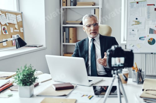 Preview: Senior man in elegant shirt and tie working using laptop while making social media video