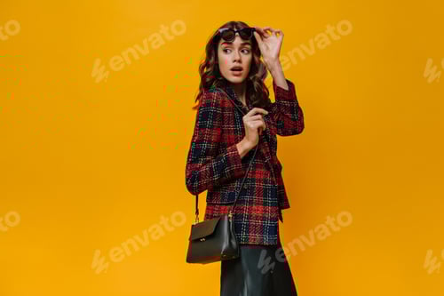 Preview: Surprised lady in red jacket and sunglasses posing on yellow backdrop. Brunette girl with cool hand
