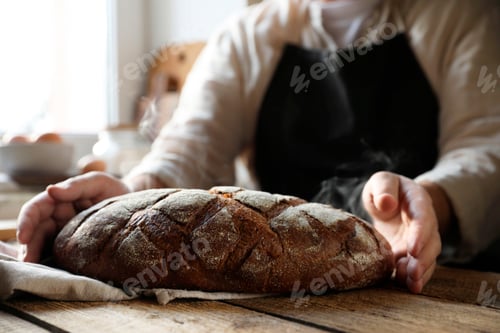 Preview: Man holding loaf of fresh bread at wooden table indoors, closeup